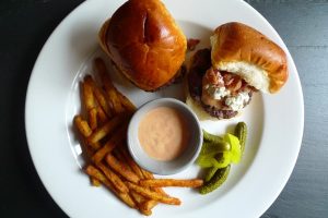 Plate showing slider-size hamburgers topped with bacon, bleu cheese, and sauce. Shown with pickles and french fries.