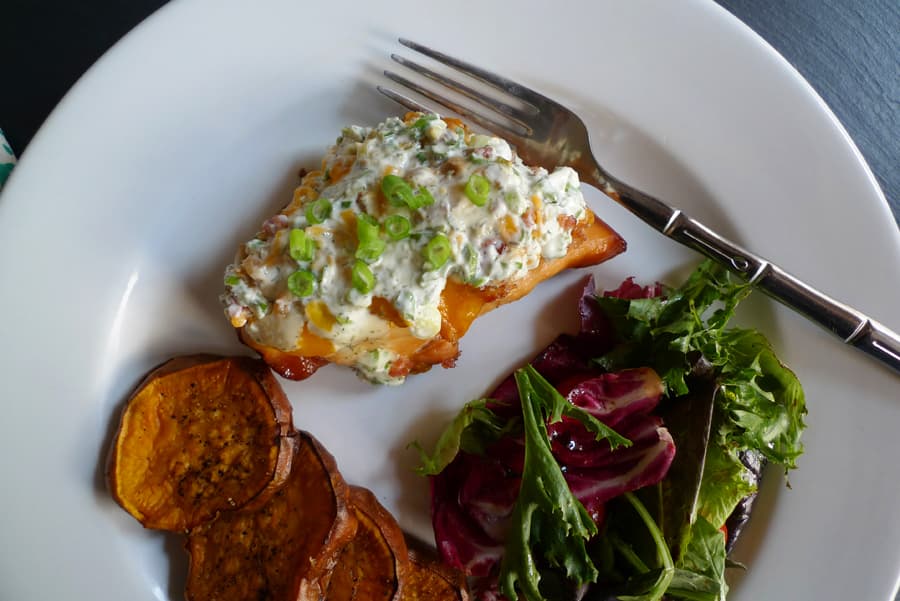Twisted Teriyaki Chicken, a teriyaki marinated chicken breast topped with cheddar, cheese, bacon, and our ranch-style sauce, shown served with salad and sweet potato slices.