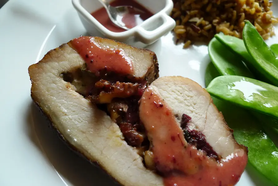 A slice of Cranberry Walnut Stuffed Pork shown plated with sauce and side dishes.